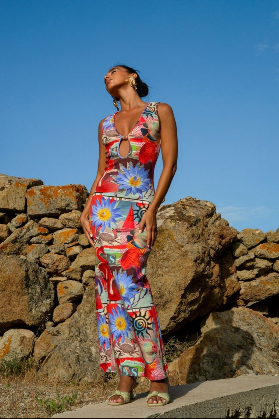 Woman in a colorful dress standing against a stone wall with a clear blue sky.